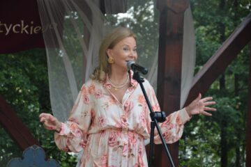Kobieta (Laura Łącz - aktorka, pisarka, reżyserka) przed mikrofonem wykonuje utwór muzyczny na scenie przy Muzeum Michała Kajki w Ogródku